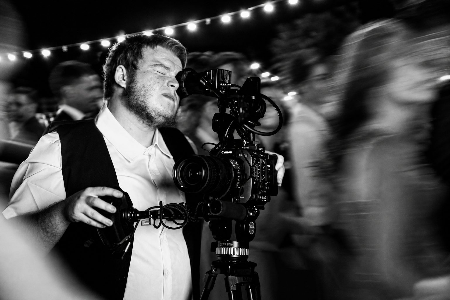 Opérateur INVprod à l'œilleton d'une caméra Canon de type cinéma sur trépied, filmant un événement en soirée éclairé de guirlandes lumineuses (photo en noir et blanc).