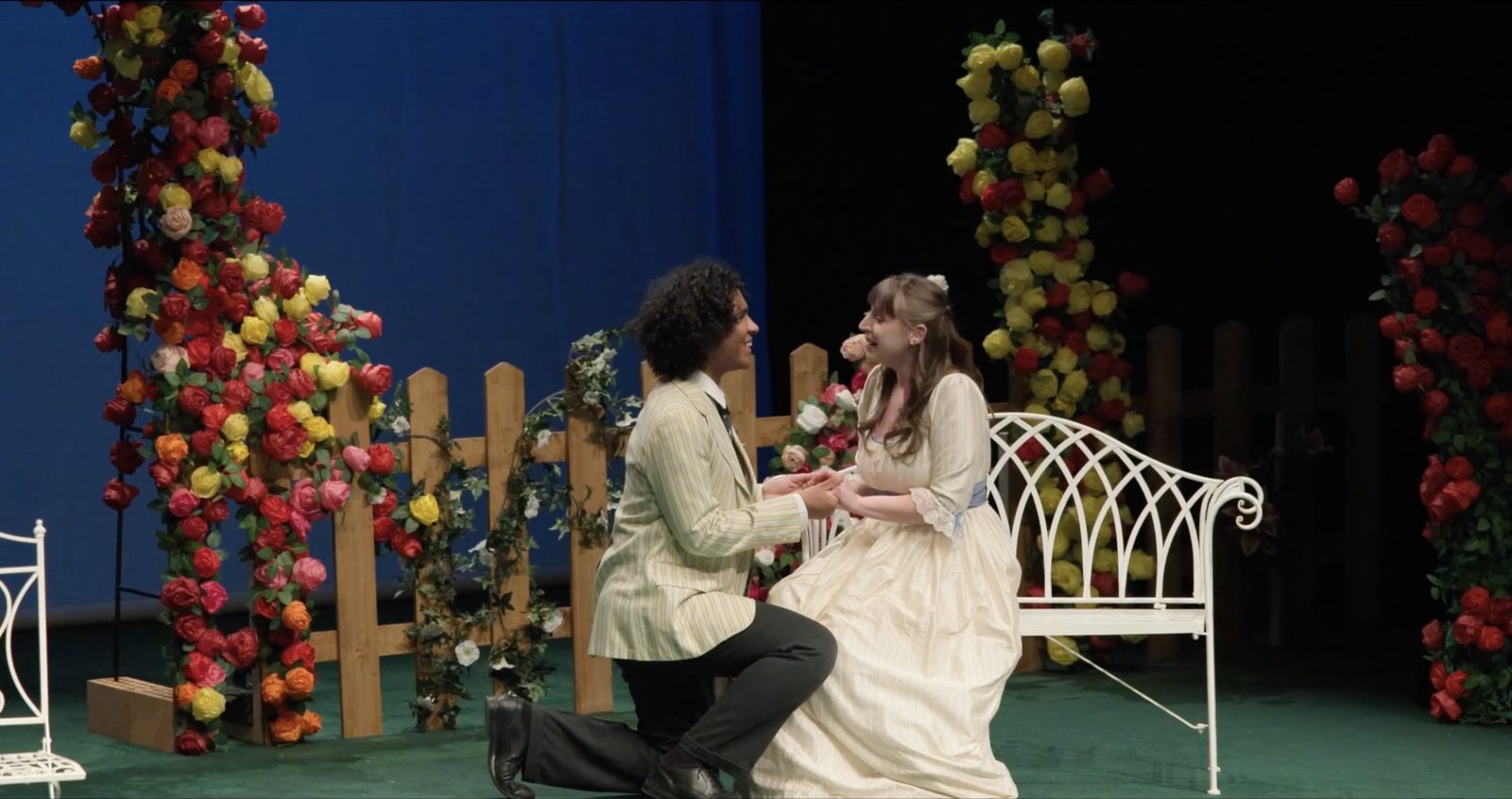 Scène de théâtre : un comédien en costume met un genou à terre devant une comédienne assise sur un banc blanc, dans un décor d’arches fleuries de roses — plan issu d’une captation de spectacle.