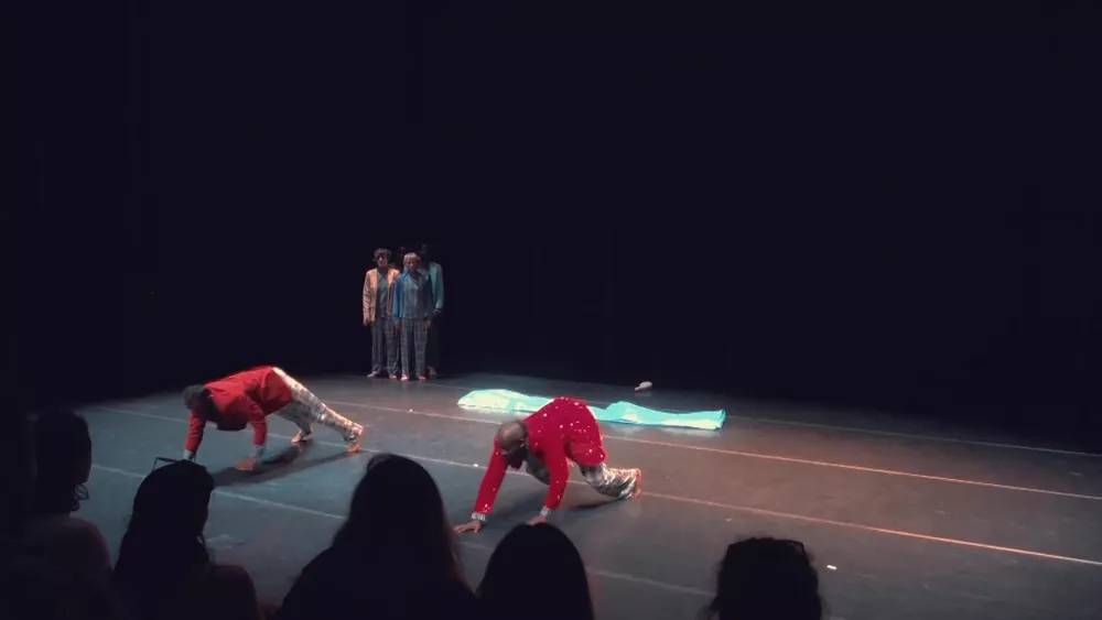 Deux danseurs en veste rouge avancent à quatre pattes, trois interprètes immobiles au fond